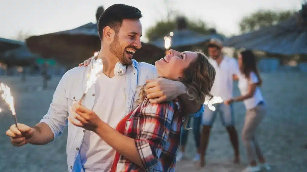 A laughing man and woman hold glowing sparklers while embracing on a sandy beach.