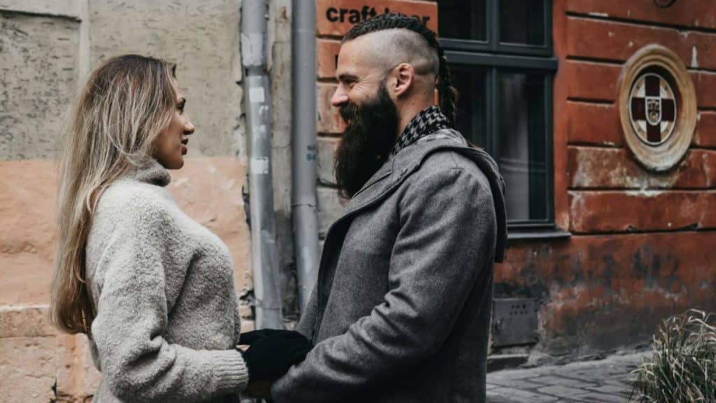 A man with a braided mohawk and long beard smiles at a woman on a street.