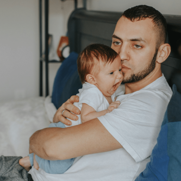 A man in a white t-shirt sits on a bed holding a yawning infant.