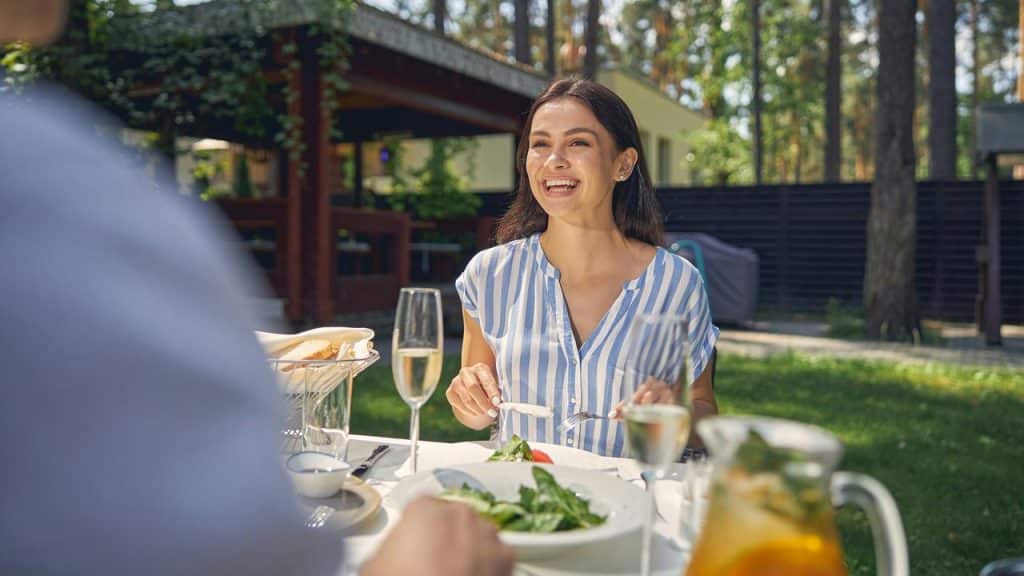 A woman smiling and enjoying a meal at an outdoor table in a sunny garden.
