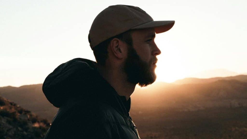 A bearded man in a cap looking toward a mountain landscape at sunset.