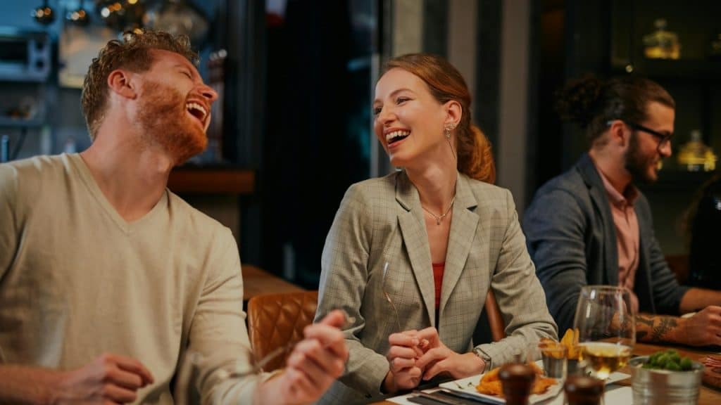 A man and woman sit at a dinner table laughing together with food in front.