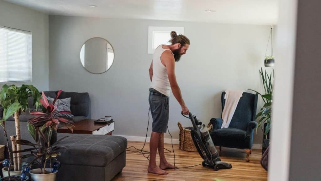 A bearded man in a tank top vacuums a wooden floor in a living room.