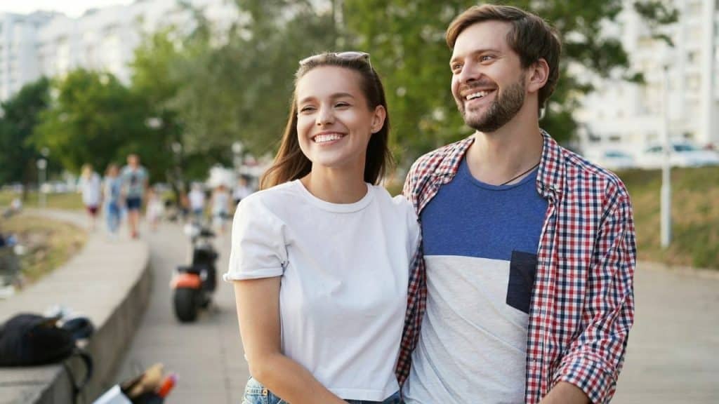 A smiling man and woman walk closely together along a paved city waterfront path.