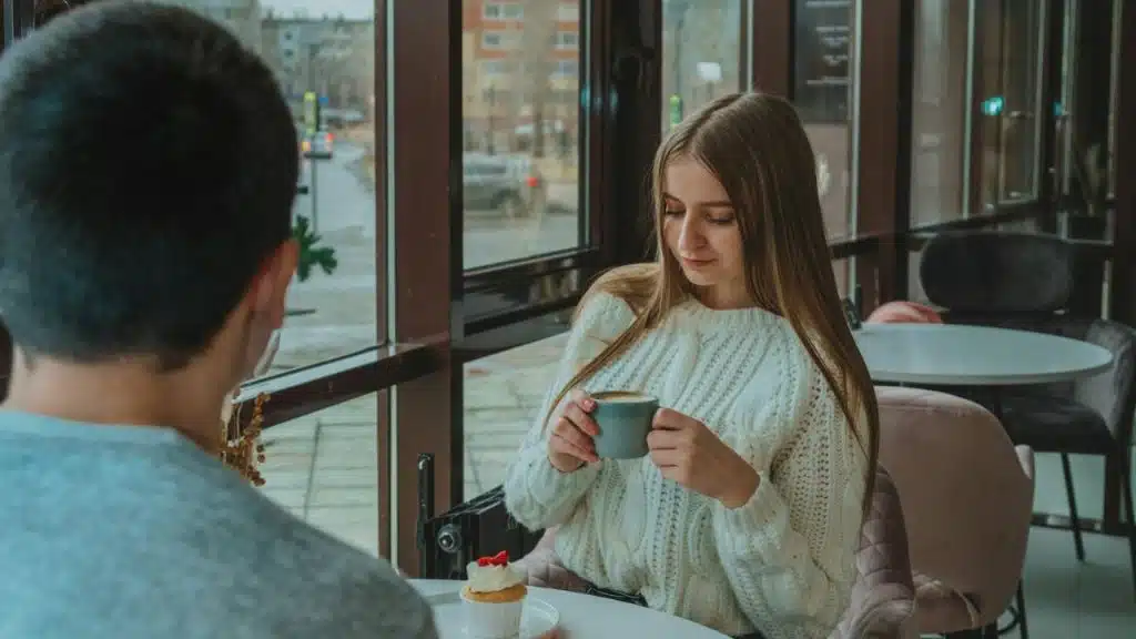 A woman in a white sweater holds a cup while sitting across from a man.
