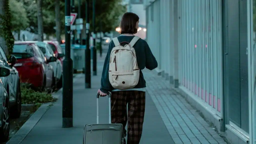 A person wearing a backpack walks down a sidewalk away from the camera, pulling a rolling suitcase.