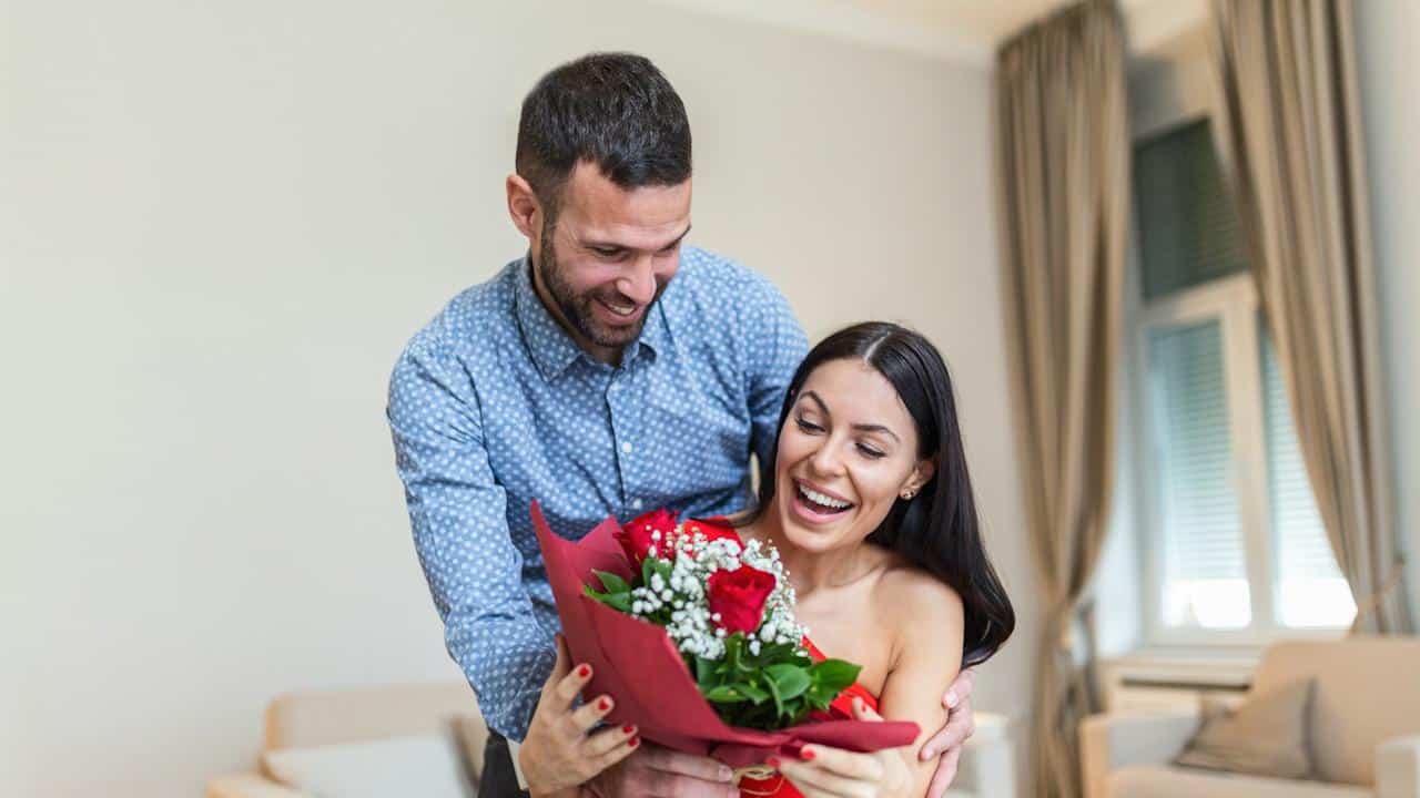 A man smiles while presenting a bouquet of red roses to a surprised and delighted woman in an indoor setting.