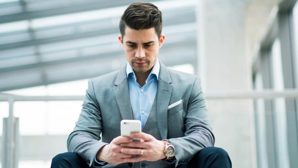 A well-dressed man sitting indoors, focused on his smartphone.