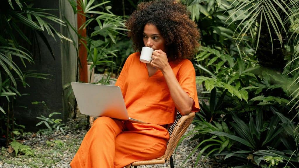 A woman sipping coffee while working on a laptop in a lush garden.