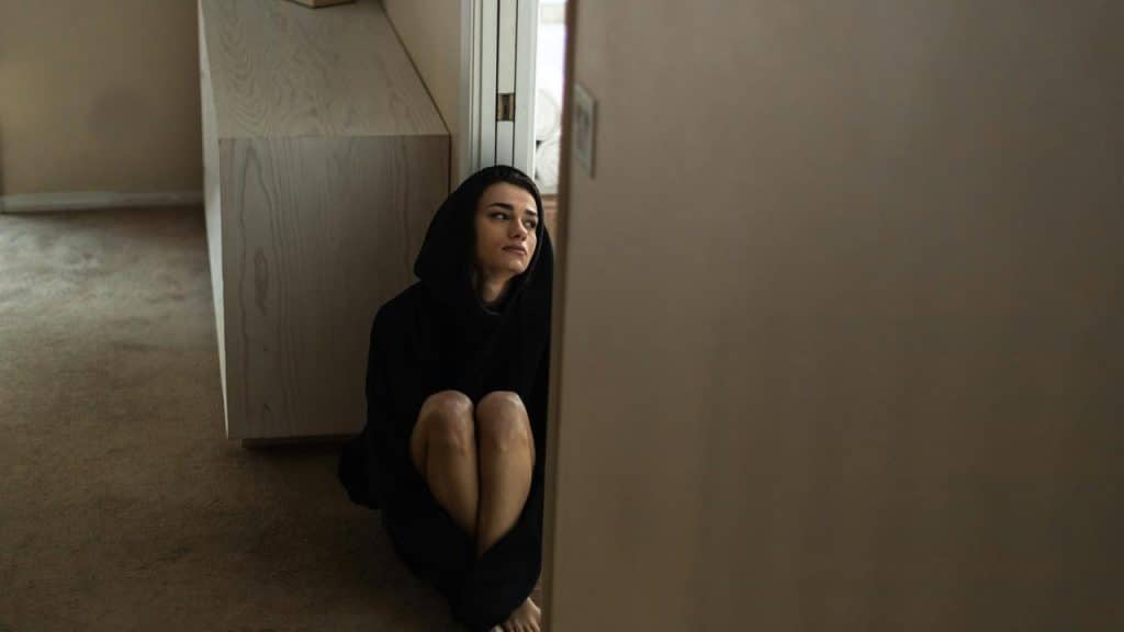 A person in a dark hoodie sitting on the floor near a doorway indoors.