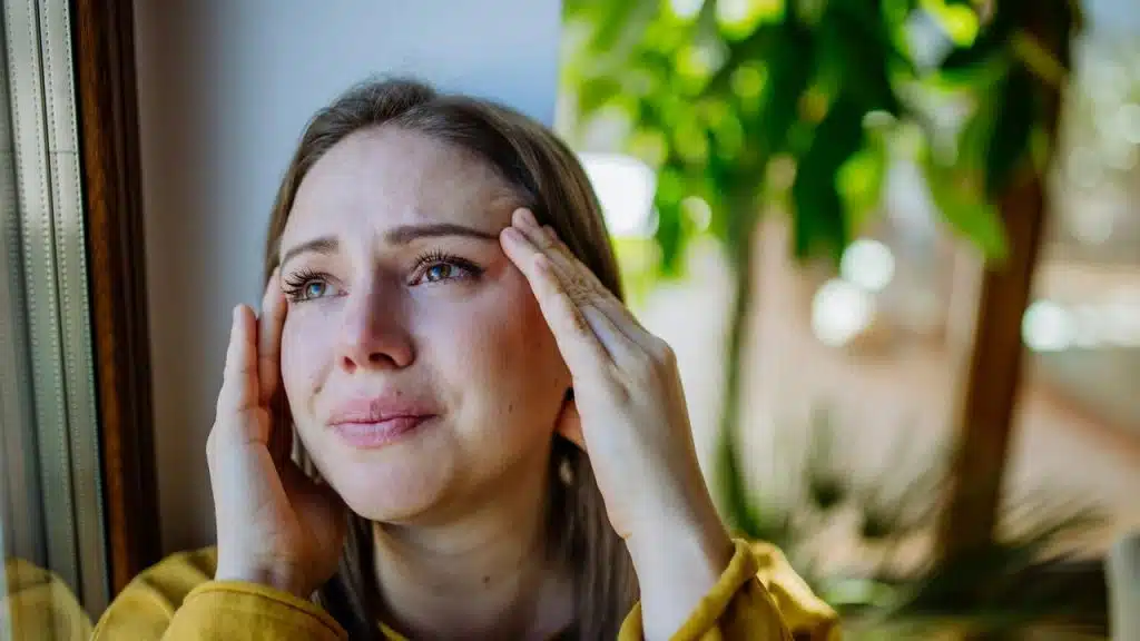 A woman holding her temples while looking distressed by a window.