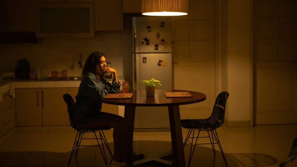A woman sits alone at a wooden table in a dimly lit kitchen under a warm pendant light.
