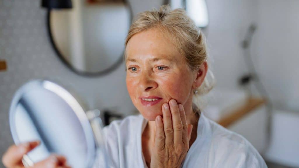 A blonde woman in a white robe looks into a handheld mirror while touching her cheek.