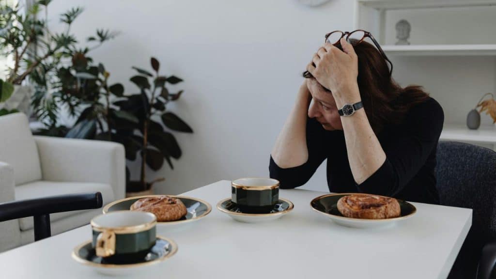 A woman sits at a table with her hands on her head, looking downward at pastries.