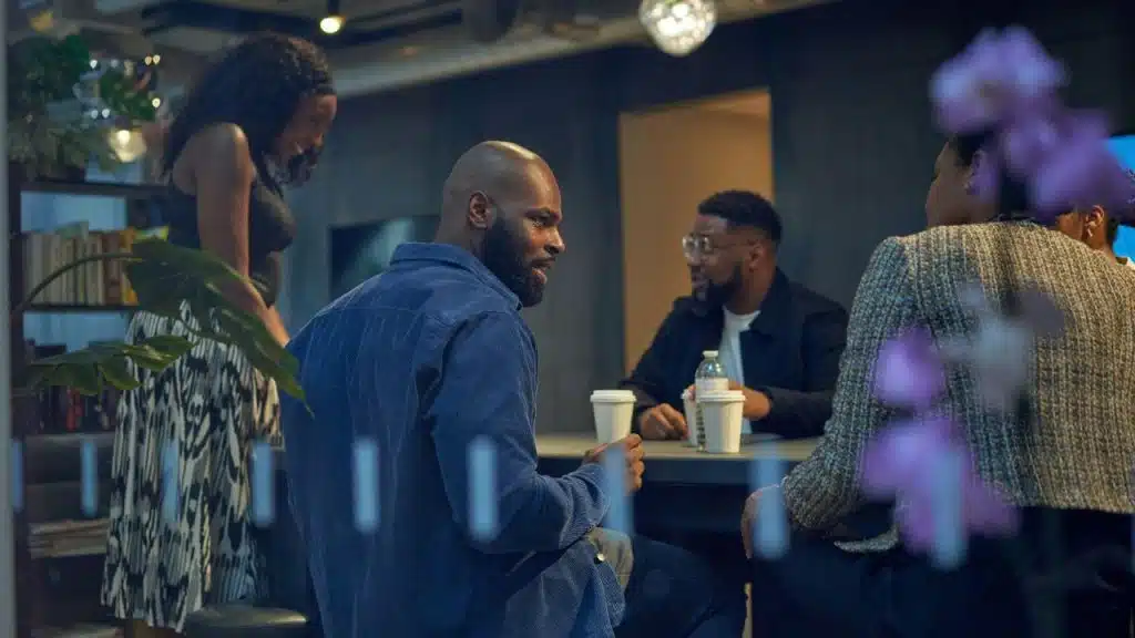 Four people sit and stand around a table in a dimly lit office, engaged in conversation.