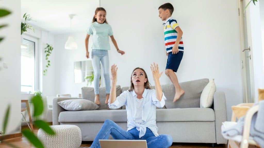 Two children jump on a sofa behind a woman sitting on the floor with arms raised.