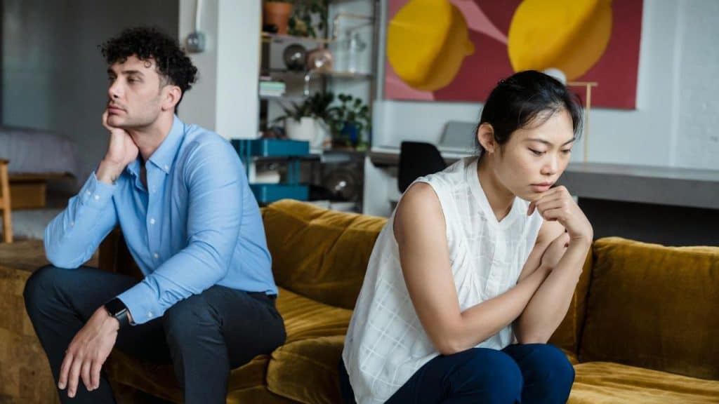 A man and woman sit back-to-back on a yellow sofa looking in opposite directions.