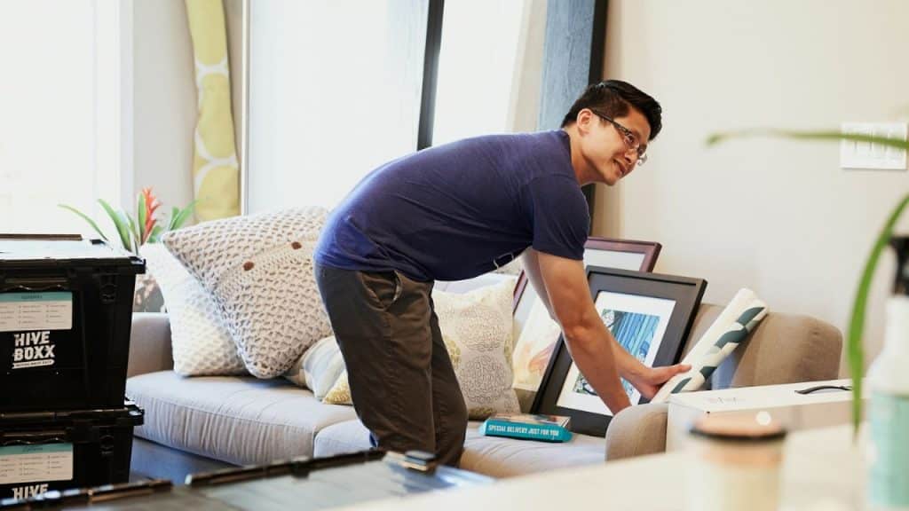 A man in a blue t-shirt and glasses leans over a couch to organize framed artwork.