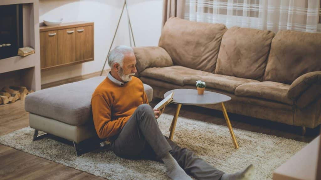 An older man in an orange sweater sits on a rug while reading a book.
