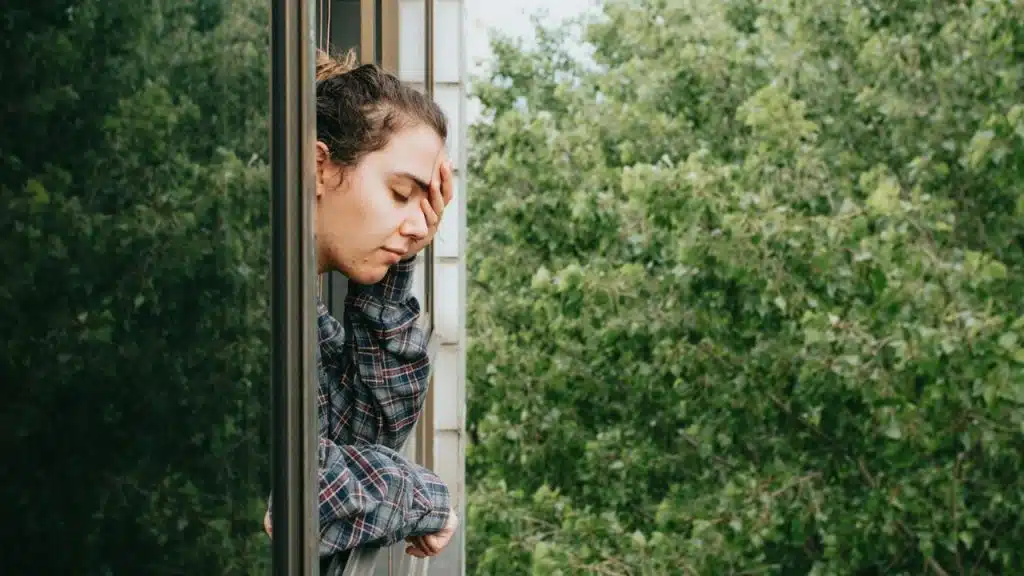 A woman in a plaid shirt rests her forehead on her hand while looking out a window.