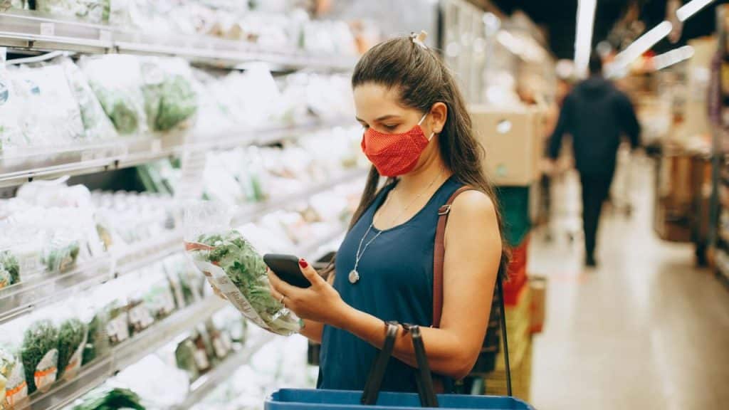 A woman wearing a red face mask holds a bag of produce and a smartphone.