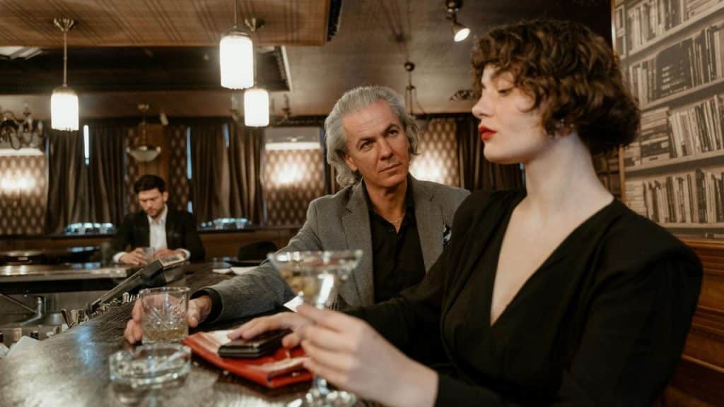 A woman in a black dress and a man in a grey jacket sit at a bar.