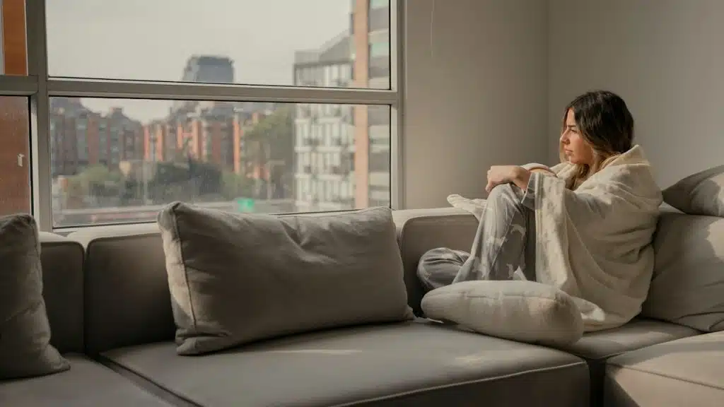 A thoughtful woman sitting on a sofa is looking out of the window.