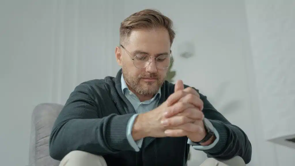 A man with glasses and a green cardigan sits with his hands clasped, looking downward.