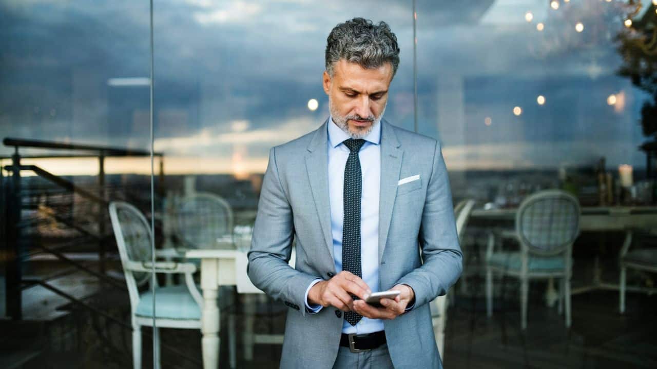 A man in a grey suit and tie stands indoors while looking down at a smartphone.