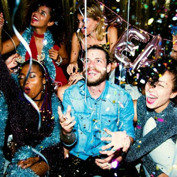 A diverse group of people celebrate together indoors while surrounded by falling colorful paper confetti.