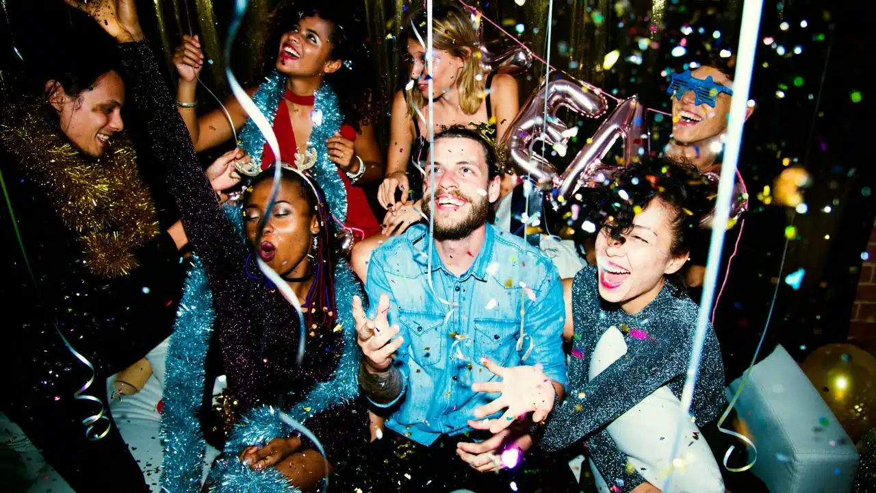 A diverse group of people celebrate together indoors while surrounded by falling colorful paper confetti.