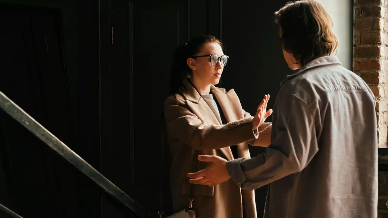A woman in a tan coat holds her hands up toward a man in a doorway.