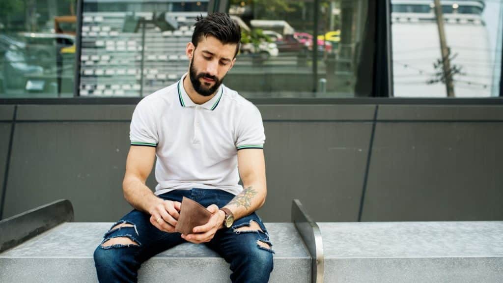 A bearded man in a white polo shirt sits outdoors and looks into a brown wallet.