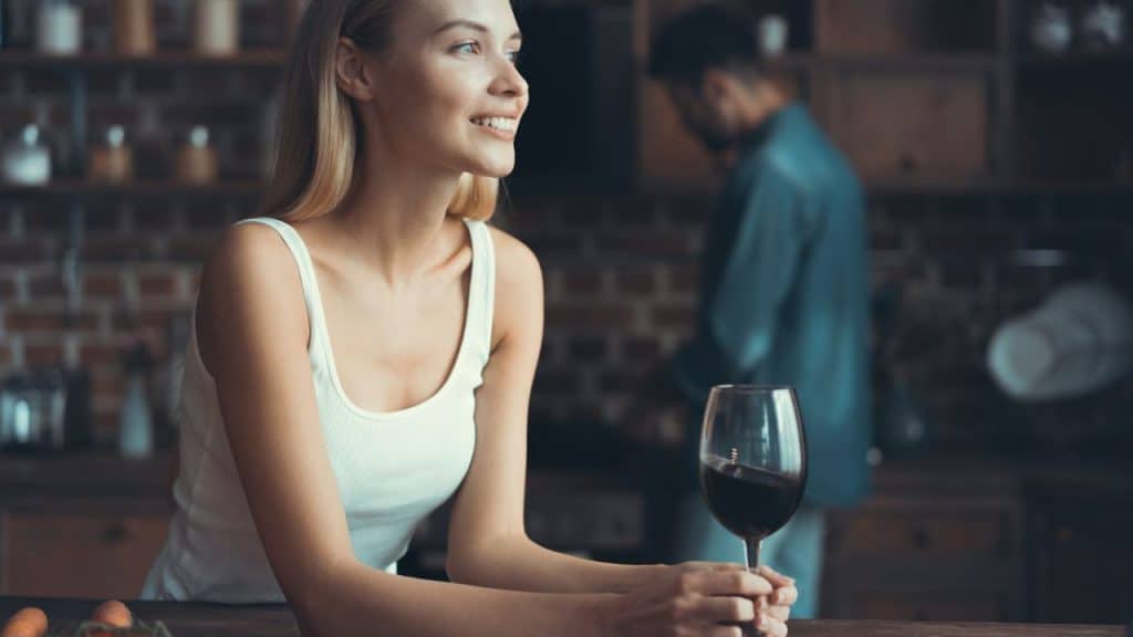 A smiling woman holds a glass of red wine while a man stands in the background.