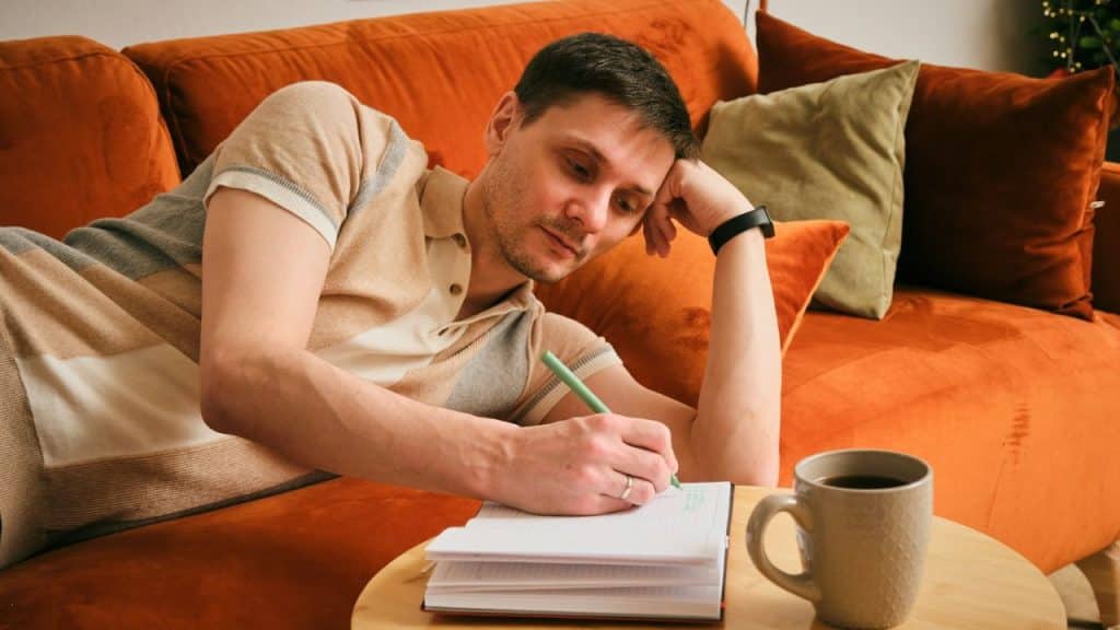 A man lies on an orange couch and writes in a notebook with a green pen.