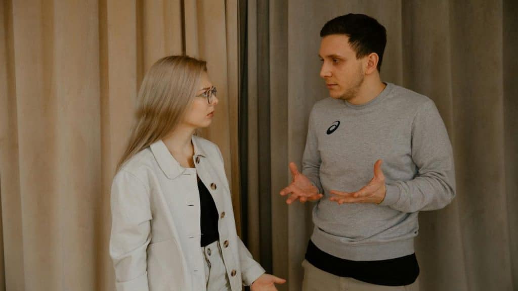 A man in a grey sweatshirt gestures with both hands while talking to a woman.