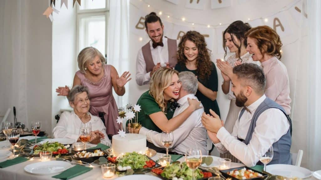A group of people at a dinner table watch a woman in green hug a man.