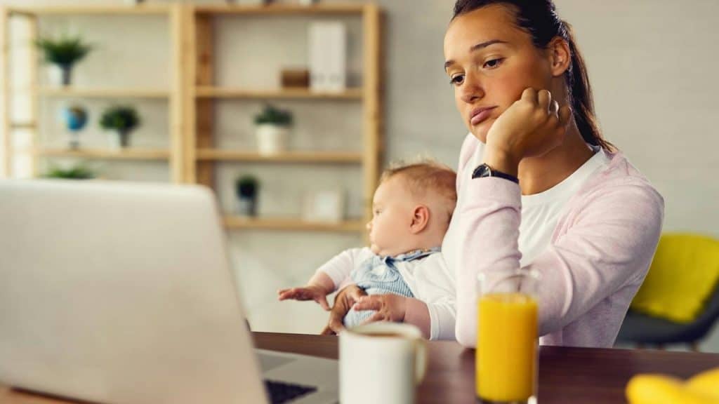 A woman holding a baby while working on a laptop at a table.