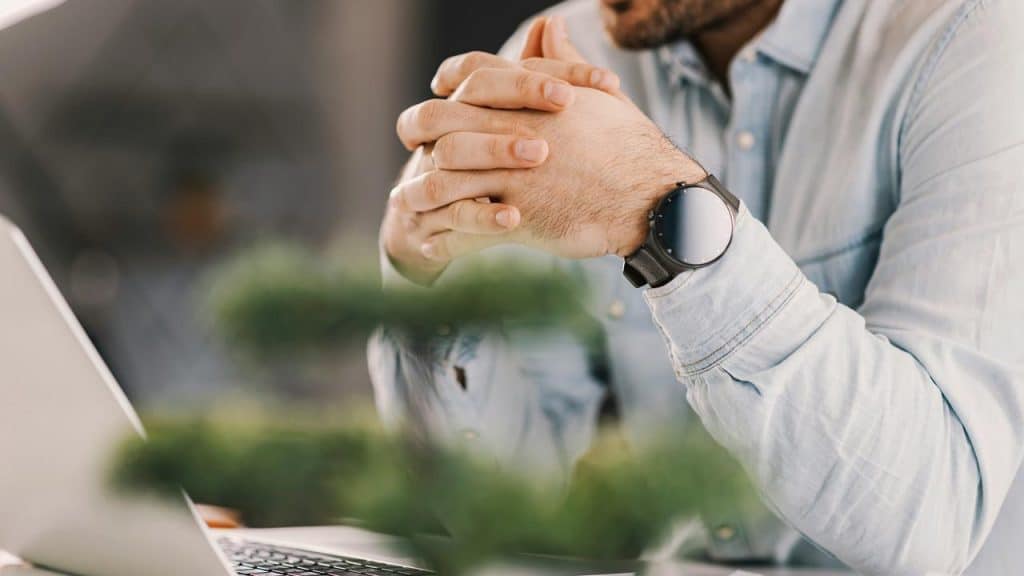 A man with clasped hands wearing a wristwatch while working on a laptop at a desk.