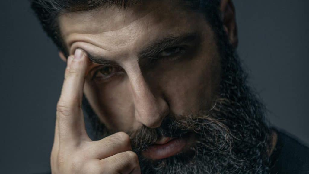 A bearded man staring at the camera with an intense expression and pushing on his eyebrow with two fingers.