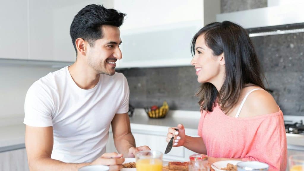 A man and woman smile at each other while eating breakfast in a bright kitchen.