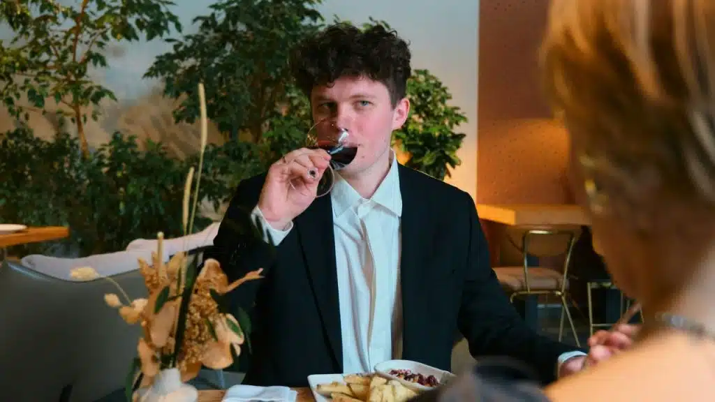 A man with curly hair in a suit drinks red wine at a table with plants.
