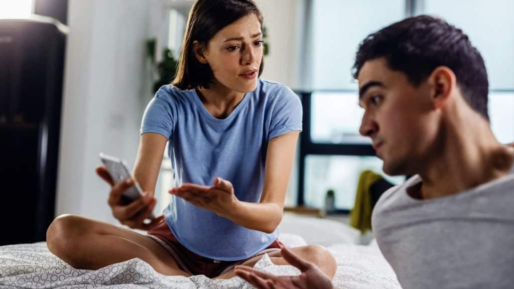 A woman holds a phone and gestures while talking to a man on a bed.