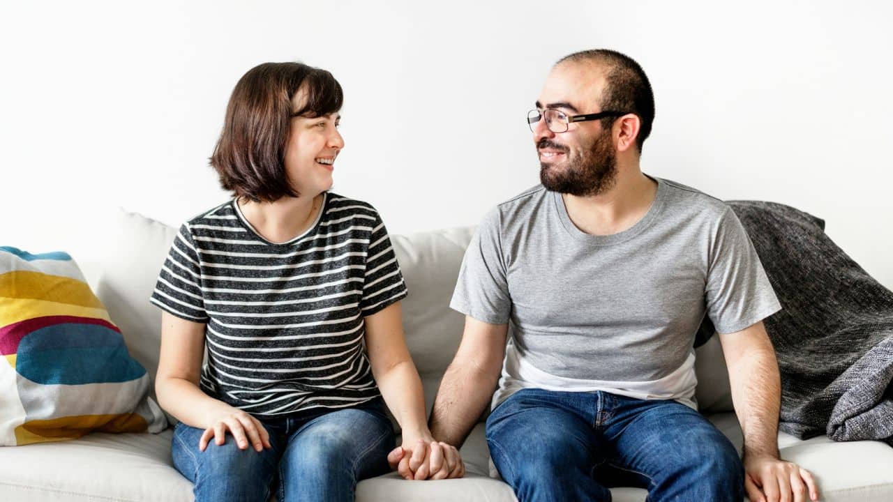 A man and woman sit on a couch holding hands and smiling at each other.