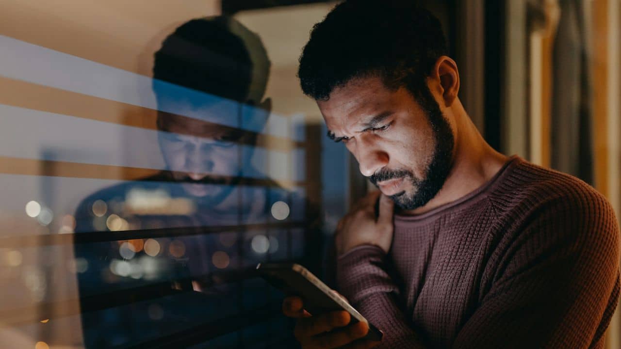 A man looks down at a smartphone while standing near a window reflecting city lights.