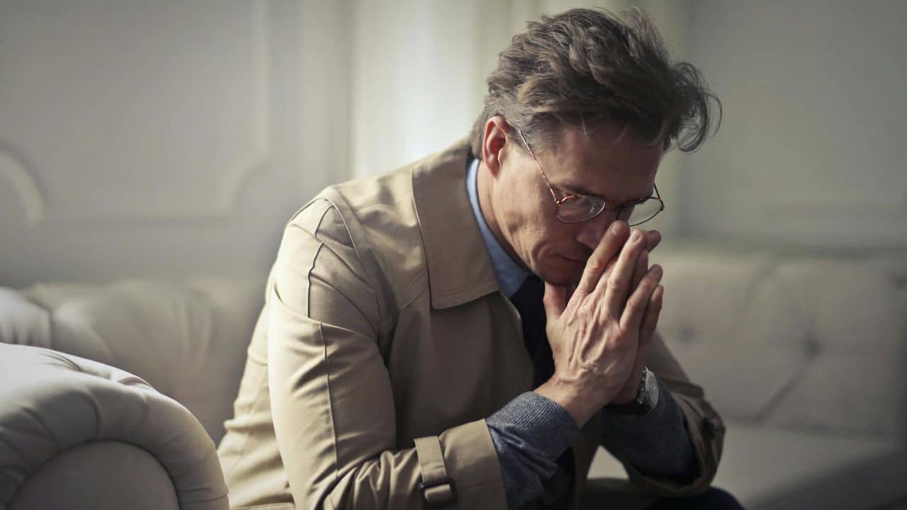 A man wearing glasses and a tan trench coat sits with his hands pressed together.