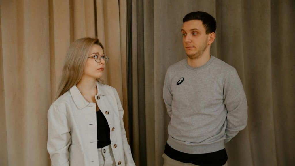 A woman with glasses and a man in a sweatshirt stand in front of tan curtains.
