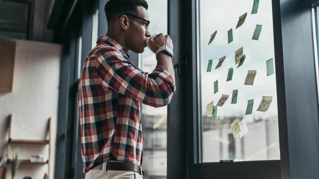 A man drinking from a cup while looking at sticky notes on a window.