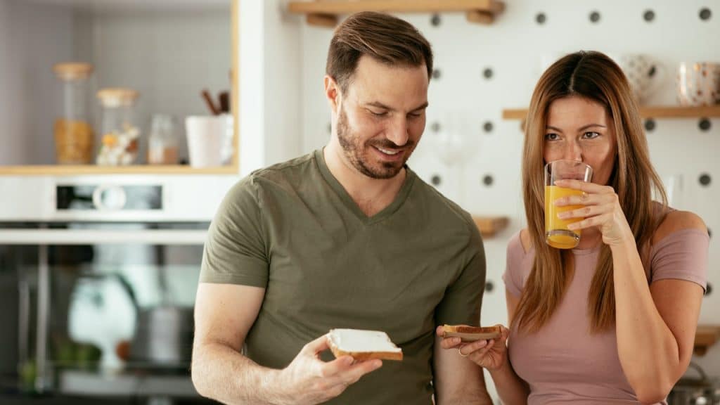 A bearded man is holding a slice of bread and smiling as a woman holds a slice in her hand and drinks juice with the other.