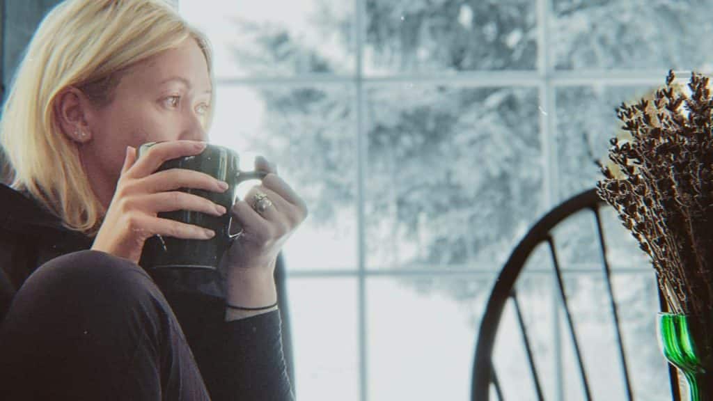 A person sitting indoors and sipping a hot drink by a window.